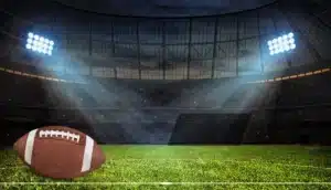 The image you provided depicts a football (specifically, American football) resting on a well-maintained grass field inside a large stadium. The stadium is illuminated by bright lights, casting an atmospheric glow that highlights the football and the surrounding area.